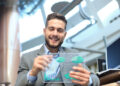 A smiling man in a suit holds a transparent futuristic screen displaying graphs and charts, illustrating the advantages of business employee benefits, as he sits in a modern, well-lit office environment. | FintechZoom