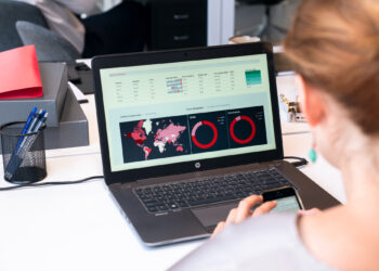 A person examines a laptop showcasing data charts and graphs, essential components of a brand awareness toolkit. The screen displays a color-coded world map alongside circle graphs labeled "Clicks" and "Conversions." A smartphone rests in their hand, surrounded by office supplies. | FintechZoom
