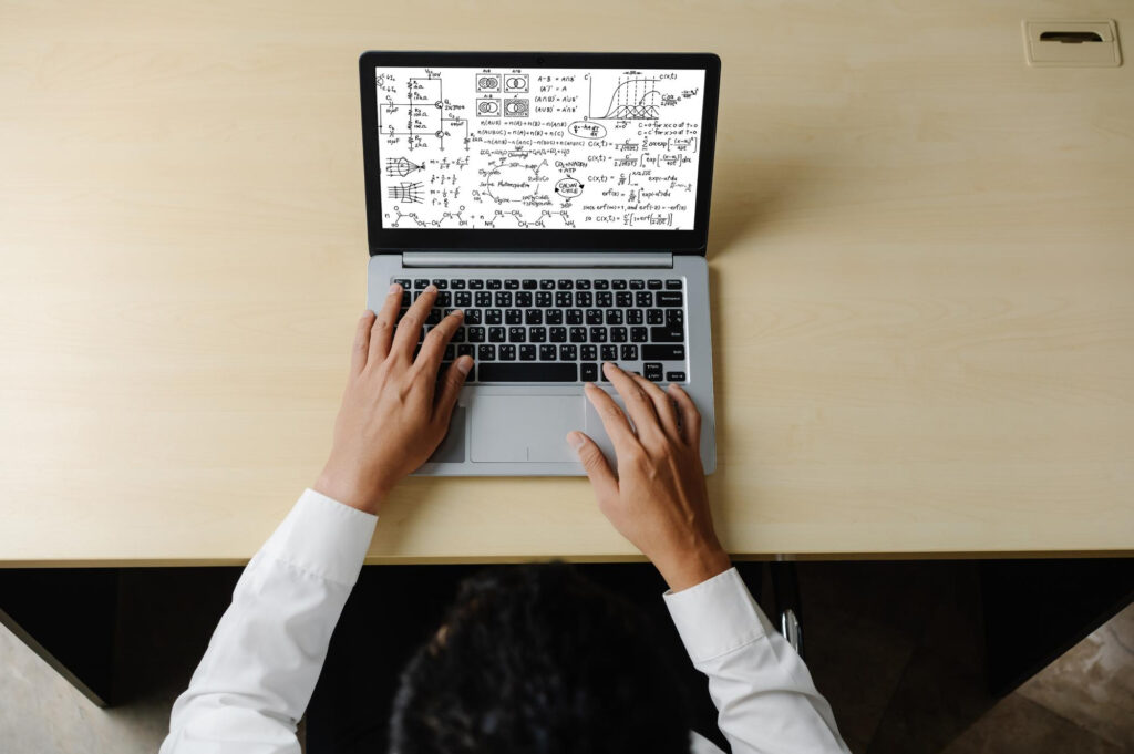 A person in a white shirt types on a laptop displaying a screen filled with complex equations and diagrams. The setting is a light wooden desk, viewed from above. | FintechZoom