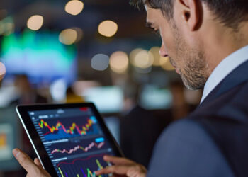 A man in a suit examines financial charts on a tablet, deeply focused on the screen that illustrates trends from the financial markets. The background is blurred with colorful lights and screens, evoking a bustling data-driven environment. | FintechZoom