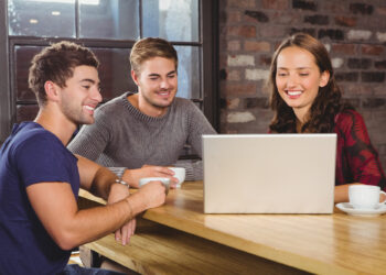 Three people sit at a wooden table, smiling as they look at a laptop screen. Two hold coffee cups, embodying how students can develop business skills. The background shows a brick wall with a window, creating an inspiring setting for collaboration and growth. | FintechZoom