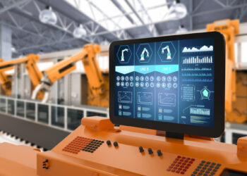 In a factory setting, robotic arms operate in the background while, upfront, a monitor showcases manufacturing data graphs labeled "Line A," "Line B," and "Line C." These are likely generated by advanced maintenance software tracking the operational status of the robotic systems. | FintechZoom