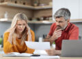 A worried couple sits at a table with a laptop, sifting through paperwork related to bad credit. The woman, in a yellow shirt, holds her head in stress, while the man, in a red polo, examines the documents thoughtfully. Books and shelves are visible in the background. | FintechZoom