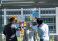 Three people in business attire stand before a glass wall, which is covered with colorful sticky notes and charts. Engaged in discussion about development teams vs. AI, they point at the notes. A cityscape is visible through the window, framing their innovative dialogue. | FintechZoom
