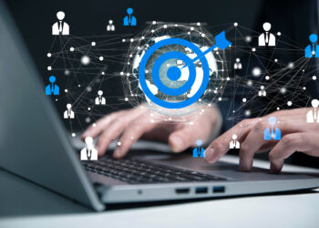Hands typing on a laptop keyboard with a digital graphic overlay. The image features a blue target symbol with an arrow, connected to icons of people, symbolizing lead generation and social connections in a networked digital landscape. | FintechZoom
