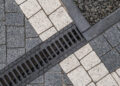A striking close-up reveals an interlocking pattern of gray and white stone pavers, seamlessly intersected by a sleek black drainage grate. This design elegantly divides a section of gravel, forming a captivating geometric composition. | FintechZoom