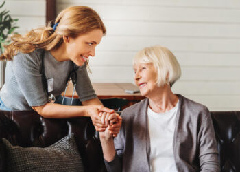 A young nurse with a stethoscope smiles as she holds hands with an elderly woman, ensuring her loved one feels safe and cherished. Both appear happy and engaged in a caring environment, set against light-colored walls and a lush plant. | FintechZoom