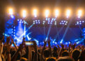 A vibrant concert scene with a large crowd under bright stage lights. People have their hands raised, one recording on a smartphone, while others discreetly enjoy nicotine pouches. The atmosphere is energetic and lively, capturing the essence of music festivals. | FintechZoom
