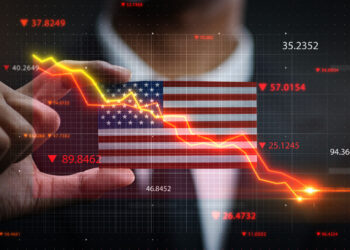 A person points at a translucent screen showing a downward-trending financial graph overlaid on the U.S. flag. Red numbers and arrows highlight declining data points, signaling a financial downturn. | FintechZoom