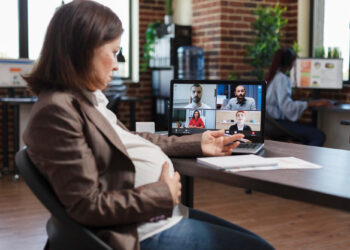 A pregnant woman sits at a desk in an office, touching her belly and looking at a laptop displaying a video call with four colleagues. Embracing remote work, she connects effortlessly with a global team. The room features brick walls, large windows, and a few plants. | FintechZoom