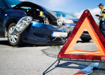 A damaged blue car involved in a collision is parked on an Atlanta road. A red warning triangle is placed in the foreground. A police officer stands in the background, examining the scene of one of many Atlanta accidents. The sky is clear and sunny. | FintechZoom