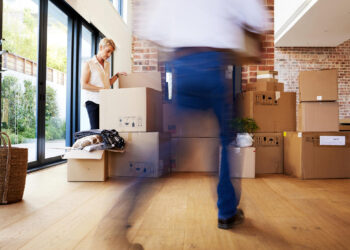 In a bright room with large windows, filled with stacked cardboard boxes, the dynamic scene reflects how the moving industry shapes real estate investing. A person blurred in motion carries a box across the light wooden floor, while a basket rests by the brick wall. | FintechZoom