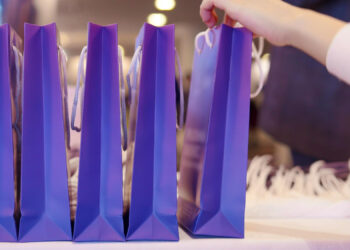 A row of upright purple custom gift bags sits neatly on a table, their white rope handles gleaming. A person reaches for one, adding a touch of anticipation. The softly blurred background hints at an indoor setting filled with excitement. | FintechZoom