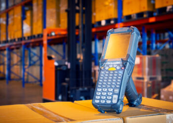 A handheld barcode scanner with a keypad rests on a stack of cardboard boxes in a warehouse. This essential piece of equipment is central to efficient inventory management, with shelves stocked full and a forklift hinting at the bustling industrial activity all around. | FintechZoom