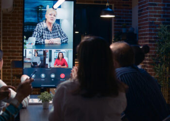 A group of people in an office watches a video call on a large screen, displaying three individuals in separate virtual windows. Perfect for hosting virtual events for companies in LATAM, the room boasts brick walls and a modern setup with various tech equipment. | FintechZoom
