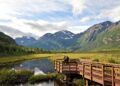 A wooden boardwalk extends over a calm stream, surrounded by Nebraska's lush greenery. Snow-capped mountains tower in the background under a partly cloudy sky. A person stands at the end of the boardwalk, soaking in the scenic view. | FintechZoom