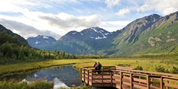 A wooden boardwalk extends over a calm stream, surrounded by Nebraska's lush greenery. Snow-capped mountains tower in the background under a partly cloudy sky. A person stands at the end of the boardwalk, soaking in the scenic view. | FintechZoom