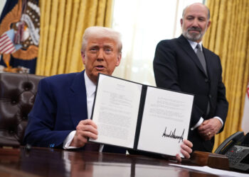 Trump holds up a signed document in front of a desk inside an office with golden curtains, symbolizing the impact of Trump tariffs. Another man stands nearby, also in a suit. | FintechZoom
