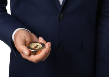 A person in a dark blue suit holds a brass compass, symbolizing the exploration of alternative investment vehicles. The white background highlights the hand prominently displaying this tool of navigation and discovery. | FintechZoom