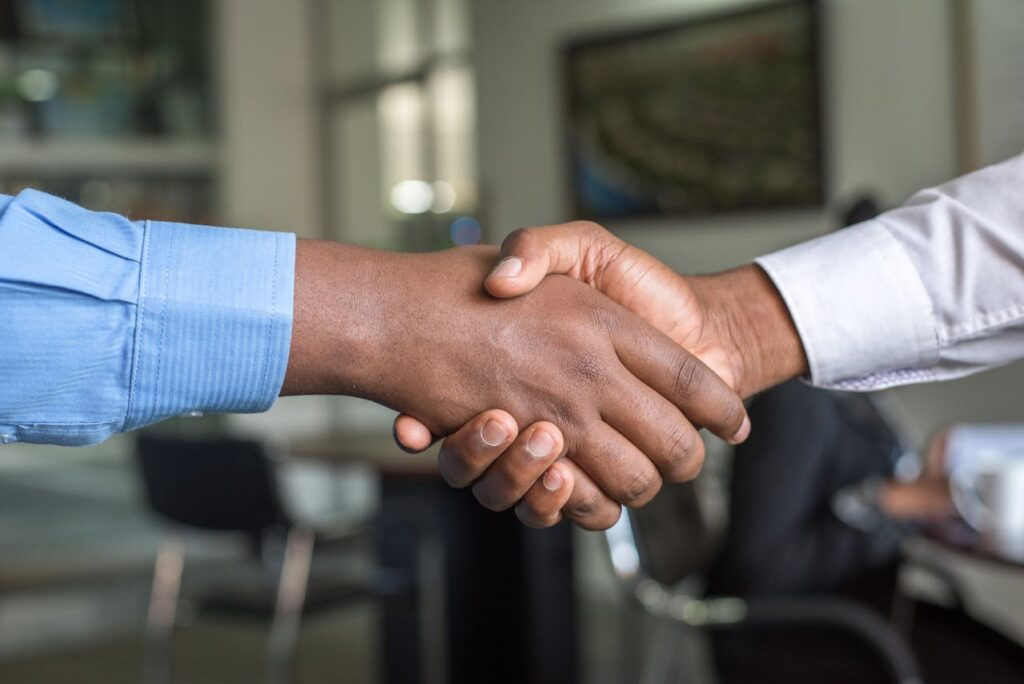 Two individuals are shaking hands in a professional setting. One person is wearing a blue shirt, and the other is in a white shirt. The background is blurred, suggesting an office environment with chairs and tables. | FintechZoom