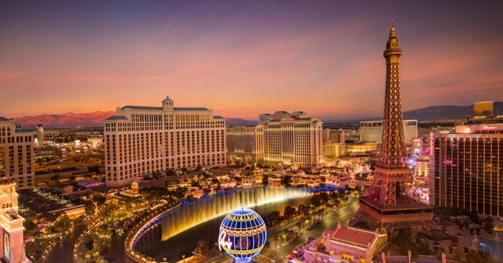 Aerial view of the Las Vegas Strip at sunset, featuring the illuminated Eiffel Tower replica, the Bellagio Hotel, and a water fountain show. The skyline is bathed in warm colors as lights begin to twinkle against the desert backdrop. | FintechZoom