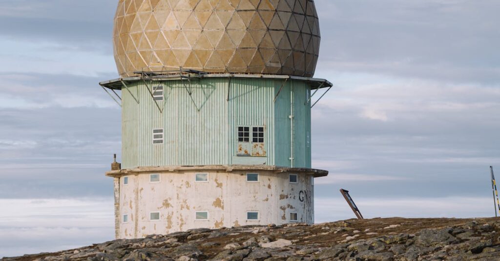 An old, abandoned radar station with a large geodesic dome sits atop a rocky hill under a cloudy sky. The building has a weathered, metal facade and various peeling paint patches. | FintechZoom