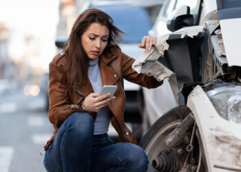 A woman in a brown jacket kneels beside a damaged car, looking worriedly at her phone. The car’s front end is badly dented, and vehicle debris is visible. The setting appears to be a city street. | FintechZoom