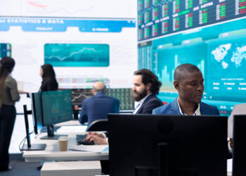 People working at computer stations in a modern office with large digital screens displaying financial data, stock charts, and a world map, highlighting the impact of automated reconciliation software on financial operations in banking. | FintechZoom