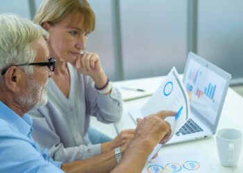 Two people are reviewing a financial report in an office. An older man with glasses points at a chart, while a woman looks on thoughtfully. They are seated at a table with papers, and a laptop displaying graphs is open in front of them, discussing how senior care franchising is a growing industry. | FintechZoom