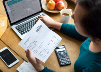 A person holding a printed invoice while sitting at a desk with a laptop, calculator, smartphone, receipts, coffee, and apples—appearing to manage finances or pay bills using invoicing and billing software. | FintechZoom
