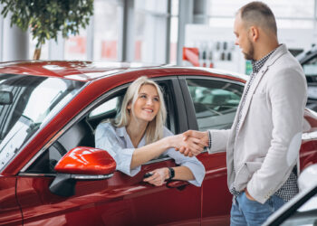 A woman sits in a red car, smiling warmly as she shakes hands with a man standing beside the vehicle. In the brightly lit showroom, the scene suggests a successful car buying experience. The man sports a light gray blazer while the woman is dressed in a light blue shirt. | FintechZoom