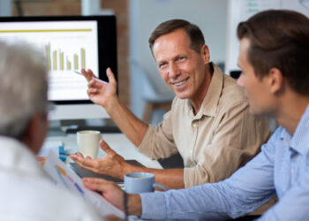 Three men in a meeting discuss data displayed on a computer monitor. One man gestures while speaking, smiling at a colleague. The atmosphere appears collaborative and focused, with mugs and documents on the table. | FintechZoom