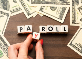A hand with red nail polish adjusts letter blocks spelling "PAYROLL" surrounded by scattered one hundred dollar bills on a wooden surface. | FintechZoom