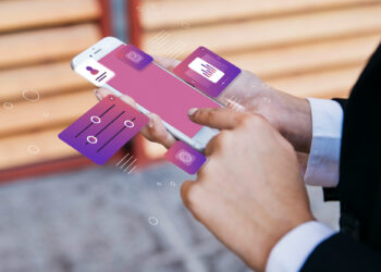 A person in business attire uses a smartphone with digital interface graphics, including charts, sliders, and icons, above the screen—suggesting advanced mobile technology like integrating Loyalty Management Software into fintech platforms. | FintechZoom