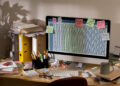 A cluttered desk with a computer displaying a spreadsheet, surrounded by papers, folders, sticky notes, pens, and a coffee mug. Sticky notes are attached to the monitor with reminders and numbers. | FintechZoom