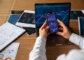 A person holds a smartphone displaying financial graphs, positioned above a laptop with similar charts on the screen. Papers with colorful data visualizations are spread on the wooden desk. | FintechZoom