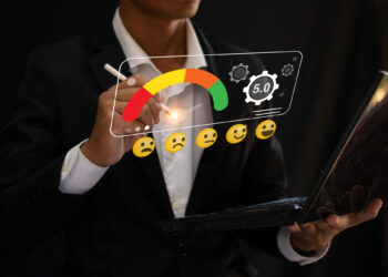 A person in a suit holds a laptop and uses a stylus to interact with a virtual customer satisfaction meter, displaying emoji faces from sad to happy and a 5.0 rating, with gears and competitor benchmarking icons in the background. | FintechZoom