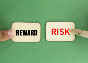 Two wooden cards on a green background—one labeled "REWARD" in black, pressed by a finger, the other labeled "RISK" in red, pointed at by a tool—show how blackjack and financial planning have something big in common. | FintechZoom