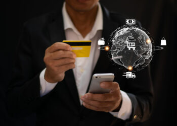 A person in a suit holds a credit card and smartphone. Nearby, a digital globe floats with shopping icons, symbolizing global e-commerce seamlessly integrated with local payment options for online transactions. | FintechZoom