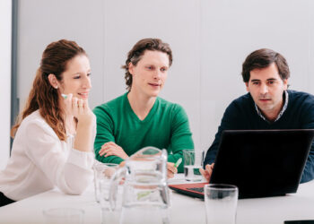Three people sit around a table with a laptop, immersed in discussing interview questions every fintech startup should ask. A woman in a white blouse and two men, one in a green sweater and the other in dark attire, focus on the screen. A glass pitcher and water glasses rest on the table. | FintechZoom
