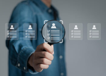 A person in a denim shirt holds a magnifying glass, focusing on virtual icons of resumes or profiles with checkmarks, symbolizing the hiring process and candidate selection in recruitment. | FintechZoom
