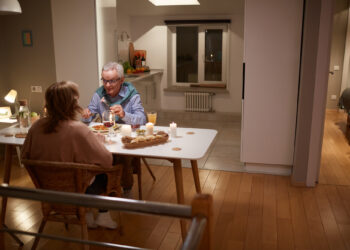 An older couple is sitting at a dining table in a cozy, warmly lit home, eating dinner together. Candles and food are on the table, and the kitchen and a bedroom are visible in the background. | FintechZoom