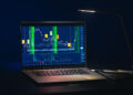 A laptop on a dark desk displays colorful financial charts of Crypto Indexes with price fluctuations, highlighted areas, and technical indicators, all illuminated by a modern desk lamp. | FintechZoom