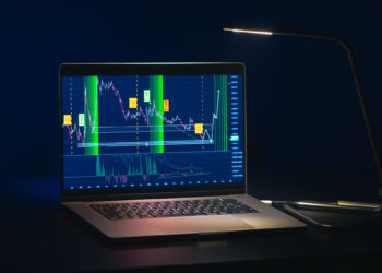 A laptop on a dark desk displays colorful financial charts of Crypto Indexes with price fluctuations, highlighted areas, and technical indicators, all illuminated by a modern desk lamp. | FintechZoom