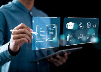 A person in a blue shirt uses a stylus to interact with virtual screens of a learning management system, displaying educational icons like a book, graduation cap, and tech symbols, while holding a tablet. The dark background glows with dynamic elements. | FintechZoom