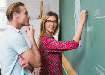 A woman in glasses writes on a chalkboard illustrated with a light bulb diagram, as a man stands beside her, contemplating. In this classroom setting featuring an easel and window, they demonstrate how to choose the best tutor for your learning style. | FintechZoom