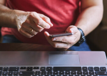 A person in a red shirt uses a smartphone with one hand while sitting at a table with an open laptop, perhaps checking instant withdrawals. The person is also wearing a watch and a wedding ring. | FintechZoom