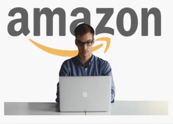 A person wearing glasses sits at a desk using a laptop, with Amazon Analytics Tools open on the screen and a large Amazon logo displayed in the background. | FintechZoom