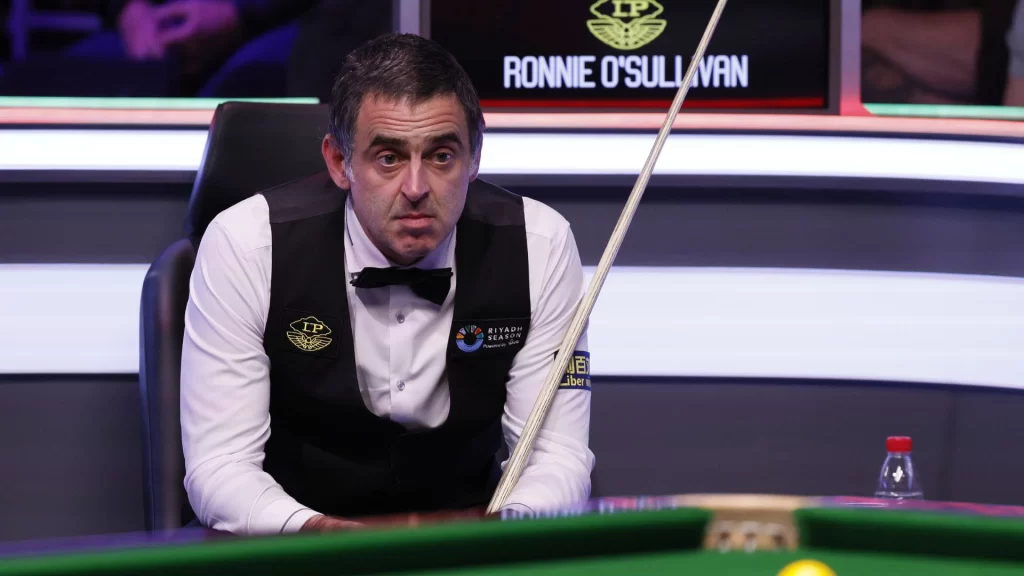 O’Sullivan professional snooker player, sits pensively by the table in a white shirt, black vest, and bow tie. His name graces the board behind him. A cue stick rests on the table beside a water bottle, capturing a moment of focused contemplation. | FintechZoom