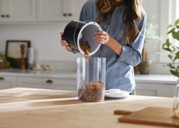A person in a denim shirt empties a container of granola or cereal into a clear canister on a wooden kitchen counter with a light, modern kitchen in the background. | FintechZoom